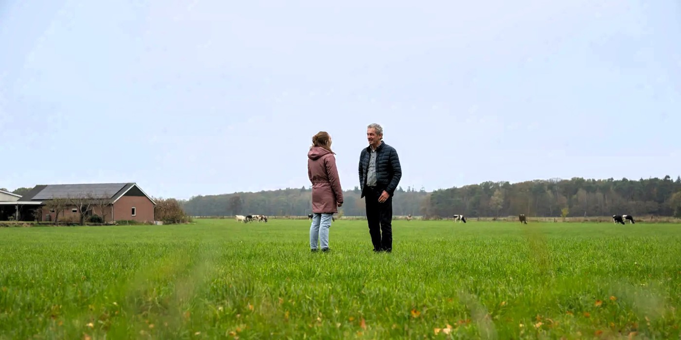 Erfcoach in gesprek met erfeigenaar in een weiland met koeien bij een boerderij. Op de achtergrond een bosrand en daarboven blauwe lucht met enkele wolken.