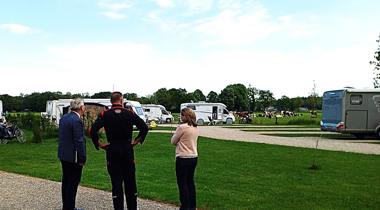 Drie mensen staan op een grindpad, omringd door geparkeerde caravans en campers op grasvelden. De lucht is deels bewolkt en er zijn bomen op de achtergrond.