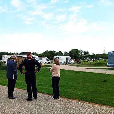 Drie mensen staan op een grindpad, omringd door geparkeerde caravans en campers op grasvelden. De lucht is deels bewolkt en er zijn bomen op de achtergrond.