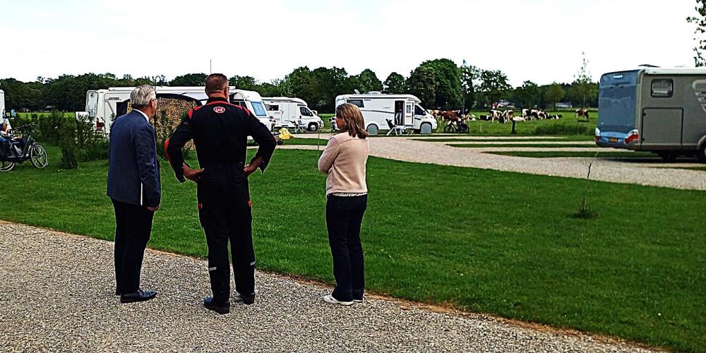 Drie mensen staan op een grindpad, omringd door geparkeerde caravans en campers op grasvelden. De lucht is deels bewolkt en er zijn bomen op de achtergrond.